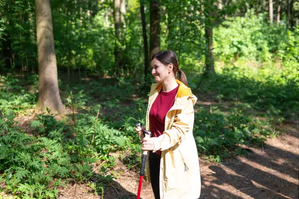 Genç bir kadın güneşli bir ormanda yürüyor, direkle İskandinav yürüyüşü yapıyor.
