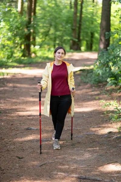 Mutlu kadın gün ışığında ormanda yürüyor, İskandinav yürüyüşü yapıyor ve dışarıda egzersiz yapıyor.