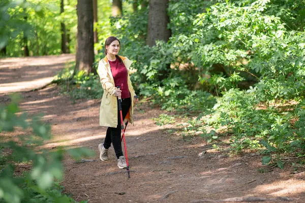 Neşeli bir kadın, güneşli bir orman patikasında sırıklarla yürüme pratiği yapıyor sağlıklı bir yaşam tarzı için zindelik ve doğayı birleştiriyor.