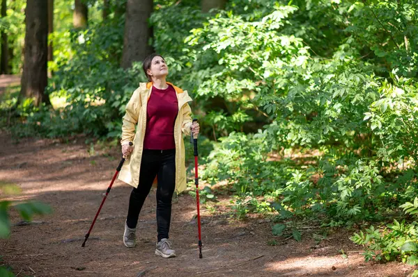 Neşeli bir kadın, güneşli bir orman patikasında sırıklarla yürüme pratiği yapıyor sağlıklı bir yaşam tarzı için zindelik ve doğayı birleştiriyor.