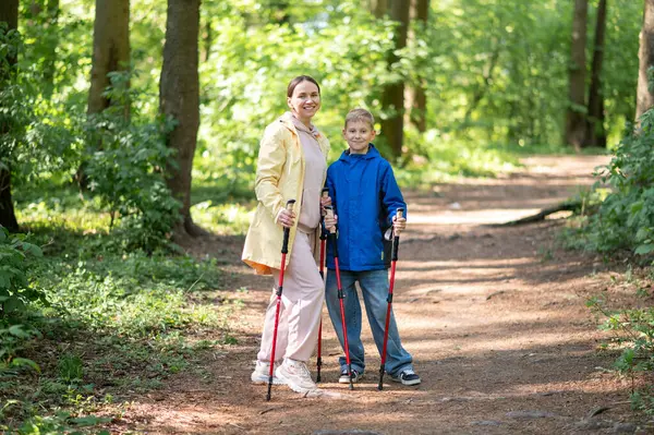 Anne ve 11 yaşındaki oğlu güneşli bir ormanda yürüyüş yapıyor ve İskandinav yürüyüşü yapıyor.
