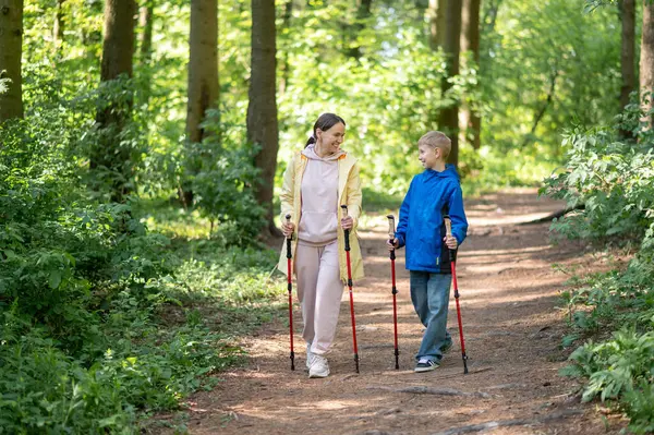 Kadın ve 11 yaşındaki oğlu, güneşli bir orman yolunda yürüyen, açık hava sporları ve aile zamanlarını kucaklayan İskandinav yürüyüşü yapıyorlar.