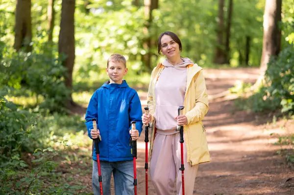 Mutlu anne ve küçük oğlu güneşli bir ormanda İskandinav direkleriyle yürüyor, doğanın ve açık hava aile kondisyonunun tadını çıkarıyor.