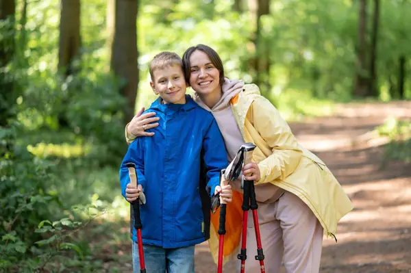 Anne, güneşli bir ormanda, bir İskandinav yürüyüşü sırasında küçük oğluna sarılıyor, sevgi dolu ve huzurlu bir açık hava anını paylaşıyor.