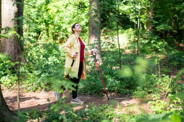 Orman yolunda özel direklerle yürüyen orta yaşlı bir kadın, sağlığı, zindeliği ve doğayla bağlantıyı teşvik ediyor.