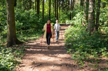 Anne ve oğlu güneşli bir ormanda yürüyor, dışarıda İskandinav yürüyüşü yapıyorlar.