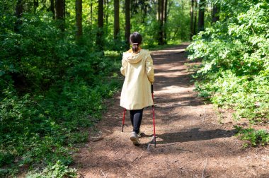 Güneşli bir günde orman yolunda yürüyen bir kadının arkadan görünüşü, doğada sırıklarla yürüme alıştırması yapıyor.