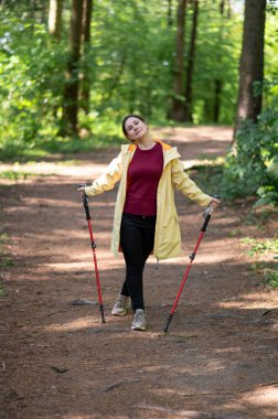Mutlu kadın gün ışığında ormanda yürüyor, İskandinav yürüyüşü yapıyor ve dışarıda egzersiz yapıyor.