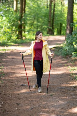 Mutlu kadın gün ışığında ormanda yürüyor, İskandinav yürüyüşü yapıyor ve dışarıda egzersiz yapıyor.