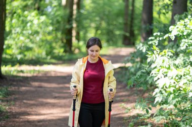 Mutlu kadın gün ışığında ormanda yürüyor, İskandinav yürüyüşü yapıyor ve dışarıda egzersiz yapıyor.