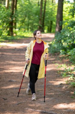 Neşeli bir kadın, güneşli bir orman patikasında sırıklarla yürüme pratiği yapıyor sağlıklı bir yaşam tarzı için zindelik ve doğayı birleştiriyor.