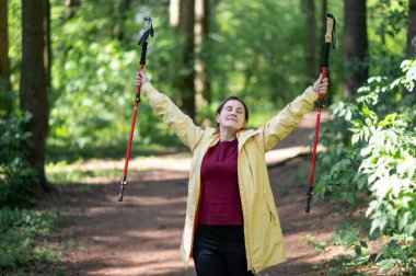 Ormandaki kadın İskandinav yürüyüş direklerini kaldırıyor, yalnızlığın ve doğayla bağın tadını çıkarıyor.