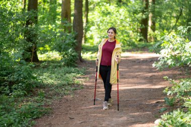 Neşeli bir kadın, güneşli bir orman patikasında sırıklarla yürüme pratiği yapıyor sağlıklı bir yaşam tarzı için zindelik ve doğayı birleştiriyor.