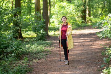 Neşeli bir kadın, güneşli bir orman patikasında sırıklarla yürüme pratiği yapıyor sağlıklı bir yaşam tarzı için zindelik ve doğayı birleştiriyor.