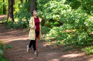 Neşeli bir kadın, güneşli bir orman patikasında sırıklarla yürüme pratiği yapıyor sağlıklı bir yaşam tarzı için zindelik ve doğayı birleştiriyor.