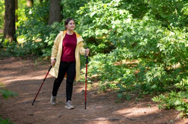 Neşeli bir kadın, güneşli bir orman patikasında sırıklarla yürüme pratiği yapıyor sağlıklı bir yaşam tarzı için zindelik ve doğayı birleştiriyor.