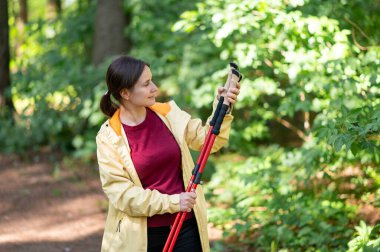 Ormanda yürüyen bir kadın, İskandinav yürüyüş direklerini inceliyor, doğanın tadını çıkarıyor ve aktif bir açık hava egzersizi için hazırlanıyor.