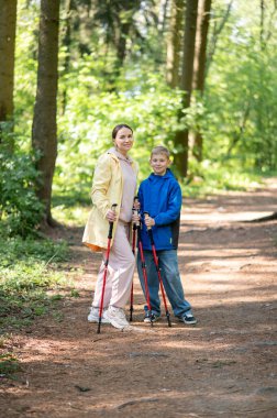 Anne ve 11 yaşındaki oğlu güneşli bir ormanda yürüyüş yapıyor ve İskandinav yürüyüşü yapıyor.