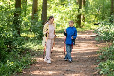 Kadın ve 11 yaşındaki oğlu, güneşli bir orman yolunda yürüyen, açık hava sporları ve aile zamanlarını kucaklayan İskandinav yürüyüşü yapıyorlar.