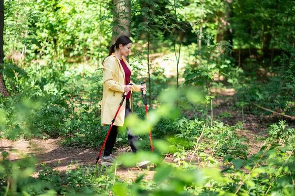 Orta yaşlı bir kadın barışçıl ormanda sırıklarla yürüyor, fiziksel egzersizle açık havada rahatlamayı birleştiriyor.