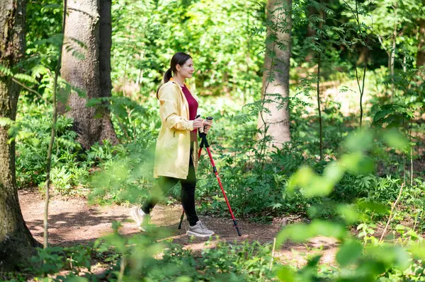 Orman yolunda özel direklerle yürüyen orta yaşlı bir kadın, sağlığı, zindeliği ve doğayla bağlantıyı teşvik ediyor.