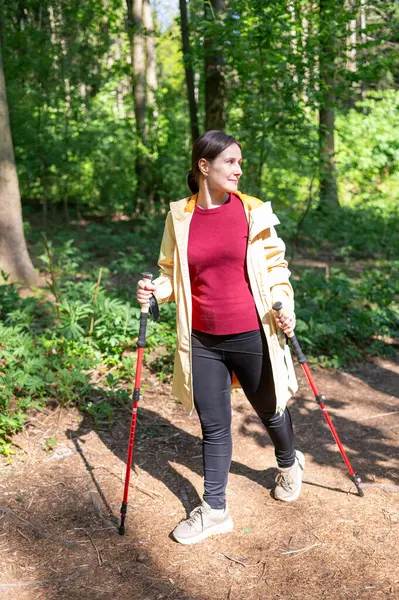 Genç bir kadın güneşli bir ormanda yürüyor, direkle İskandinav yürüyüşü yapıyor.