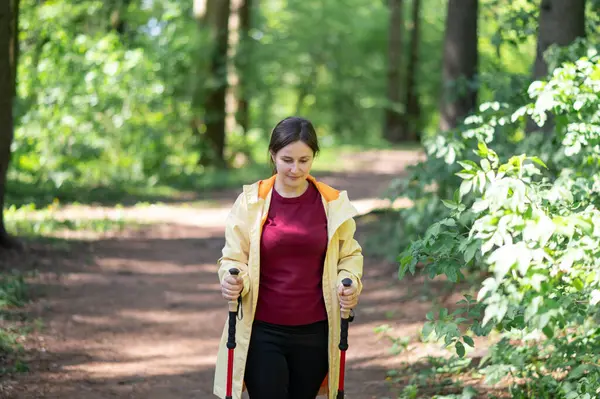 Mutlu kadın gün ışığında ormanda yürüyor, İskandinav yürüyüşü yapıyor ve dışarıda egzersiz yapıyor.