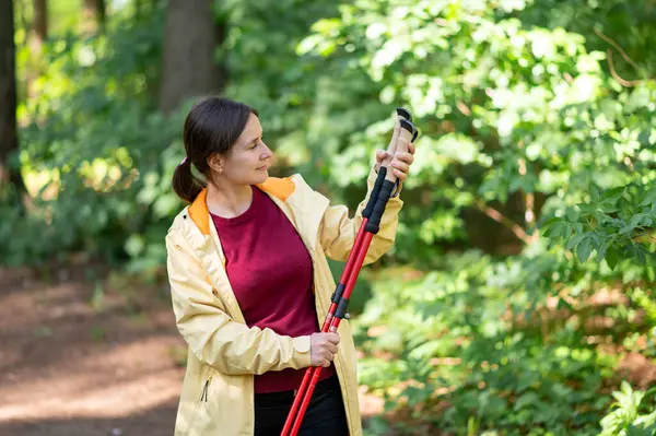 Ormanda yürüyen bir kadın, İskandinav yürüyüş direklerini inceliyor, doğanın tadını çıkarıyor ve aktif bir açık hava egzersizi için hazırlanıyor.