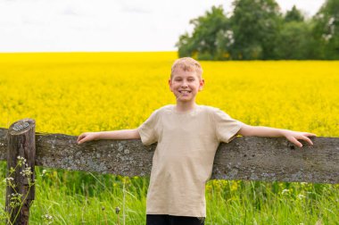 Mutlu sarışın çocuk doğada dinlenen sarı çiçek açan kolza tohumu tarlasının önündeki tahta çitin yanında yaz gününün tadını çıkarıyor.