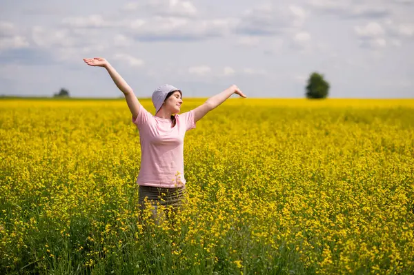 Kova şapkalı kadın kollarını kaldırıyor, sarı kolza tohumu tarlasında sıcak bir yaz gününün tadını çıkarıyor.