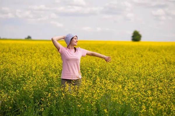 Güneş şapkalı mutlu genç kadın sıcak bir yaz öğleden sonrasında sarı kolza tohumu çiçeklerinin güzelliğine hayran.