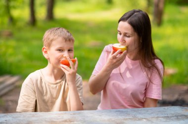 Anne ve oğul elma yiyor, gülümsüyor ve parkta meyve paylaşıyor.