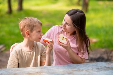 Anne ve oğul elma yiyor, gülümsüyor ve parkta meyve paylaşıyor.