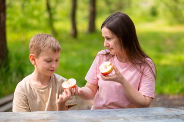Anne ve oğul elma yiyor, gülümsüyor ve parkta meyve paylaşıyor.