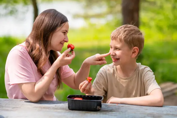 Anne ve oğlu yeşil orman parkında bir piknik masasında çilek yiyorlar.
