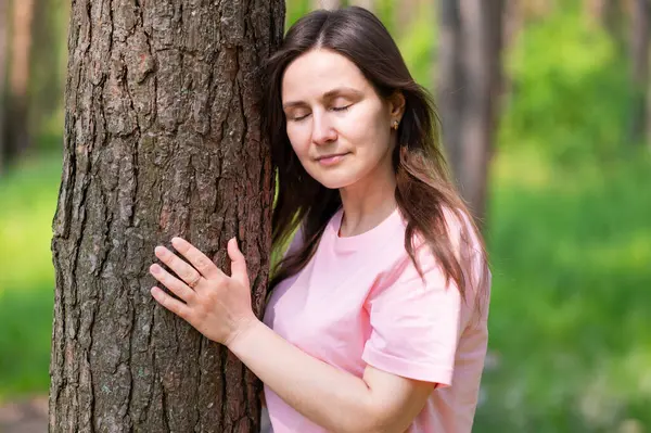 Genç bir kadın gözleri kapalı duruyor ve bir ağaca sarılıyor. Huzurlu bir orman anının tadını çıkarıyor.
