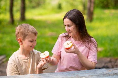 Anne ve oğul elma yiyor, gülümsüyor ve parkta meyve paylaşıyor.
