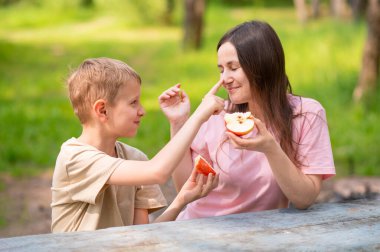 Anne ve oğul elma yiyor, gülümsüyor ve parkta meyve paylaşıyor.