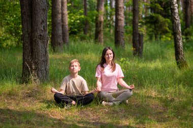 Anne ve oğul güneşli bir ormanda meditasyon yapıyorlar. Berrak, huzurlu bir aile anı.
