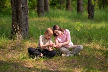 Anne ve oğlu bir ormanda yan yana oturmuş avuçlarındaki bir şeye bakıyorlar.