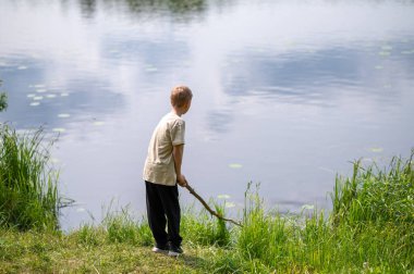 Çocuk uzun bir sopayla gölde duruyor, doğada bir yaz günü suyun yanında oynuyor.