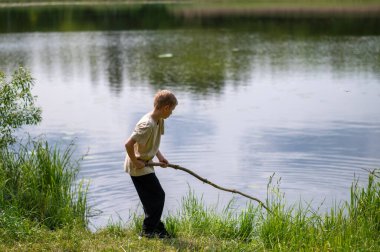 Çocuk uzun bir sopayla gölde duruyor, doğada bir yaz günü suyun yanında oynuyor.