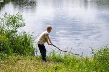 Çocuk uzun bir sopayla gölde duruyor, doğada bir yaz günü suyun yanında oynuyor.