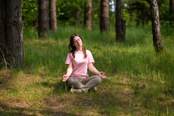 Huzurlu bir ormanda tek başına meditasyon yapan genç bir kadın, yeşil çimlerin üzerinde bağdaş kurarak oturuyor.