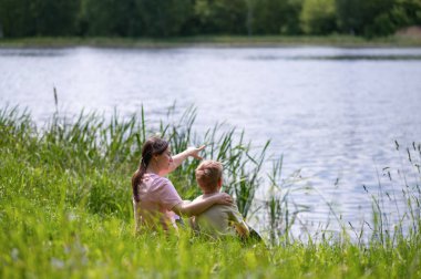 Anne ve oğul bir yaz günü gölün kenarında oturuyorlar, anne uzaklığı gösteriyor, doğada kaynaşıyorlar.