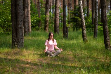 Huzurlu bir ormanda tek başına meditasyon yapan genç bir kadın, yeşil çimlerin üzerinde bağdaş kurarak oturuyor.