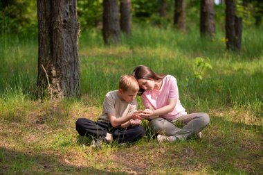 Anne ve oğlu bir ormanda yan yana oturmuş avuçlarındaki bir şeye bakıyorlar.