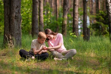 Anne ve oğlu bir ormanda yan yana oturmuş avuçlarındaki bir şeye bakıyorlar.