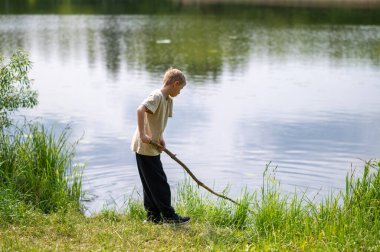 Çocuk uzun bir sopayla gölde duruyor, doğada bir yaz günü suyun yanında oynuyor.