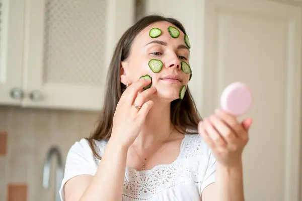 Yüzünde salatalık dilimleri olan bir kadın el aynasına bakar ve sağlıklı cilt bakımından zevk alır.